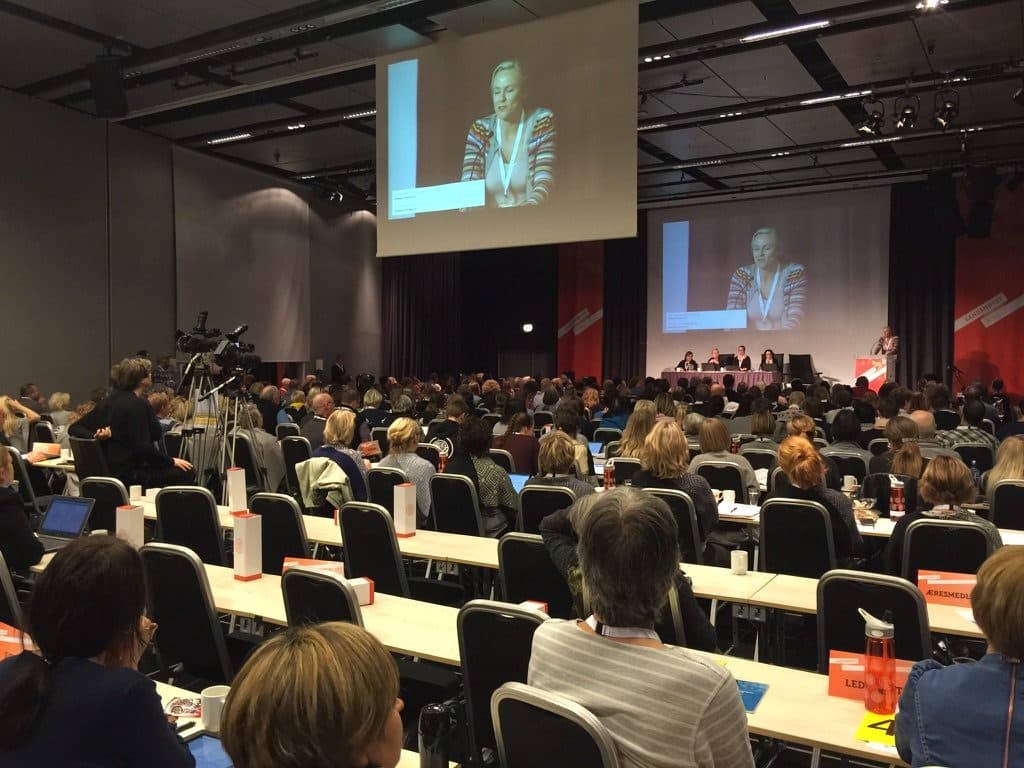 Large annual meeting seen from the hall, with table rows, a stage and screens above the audience