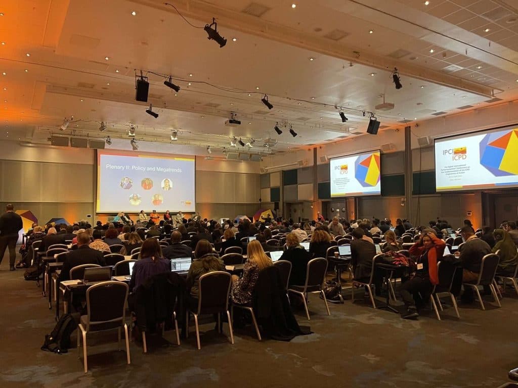 Stort plenum i en konferansesal, med deltakere ved bord og flere presentasjonsskjermer